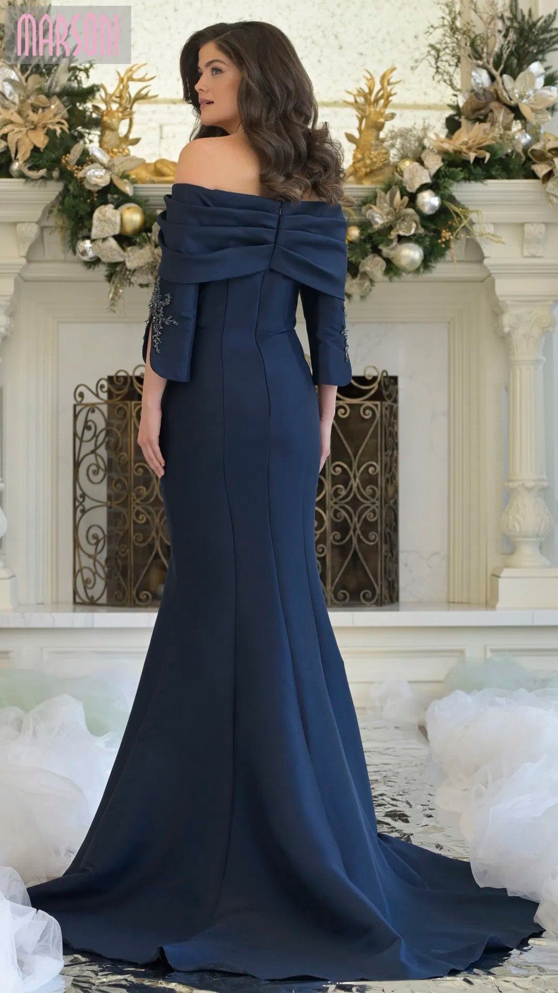 A woman stands facing away in front of a decorated white fireplace, wearing the Marsoni MV1360 long formal navy off-the-shoulder evening dress.